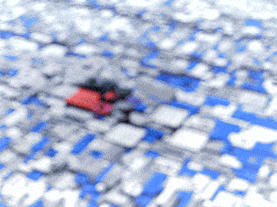 A blurry image of a cityscape reveals a matrix-like pattern of blue and white rooftops. Modular hints emerge with indistinct red and black shapes, suggesting vehicles or smaller structures. The overall scene takes on an abstract feel due to its out-of-focus quality.